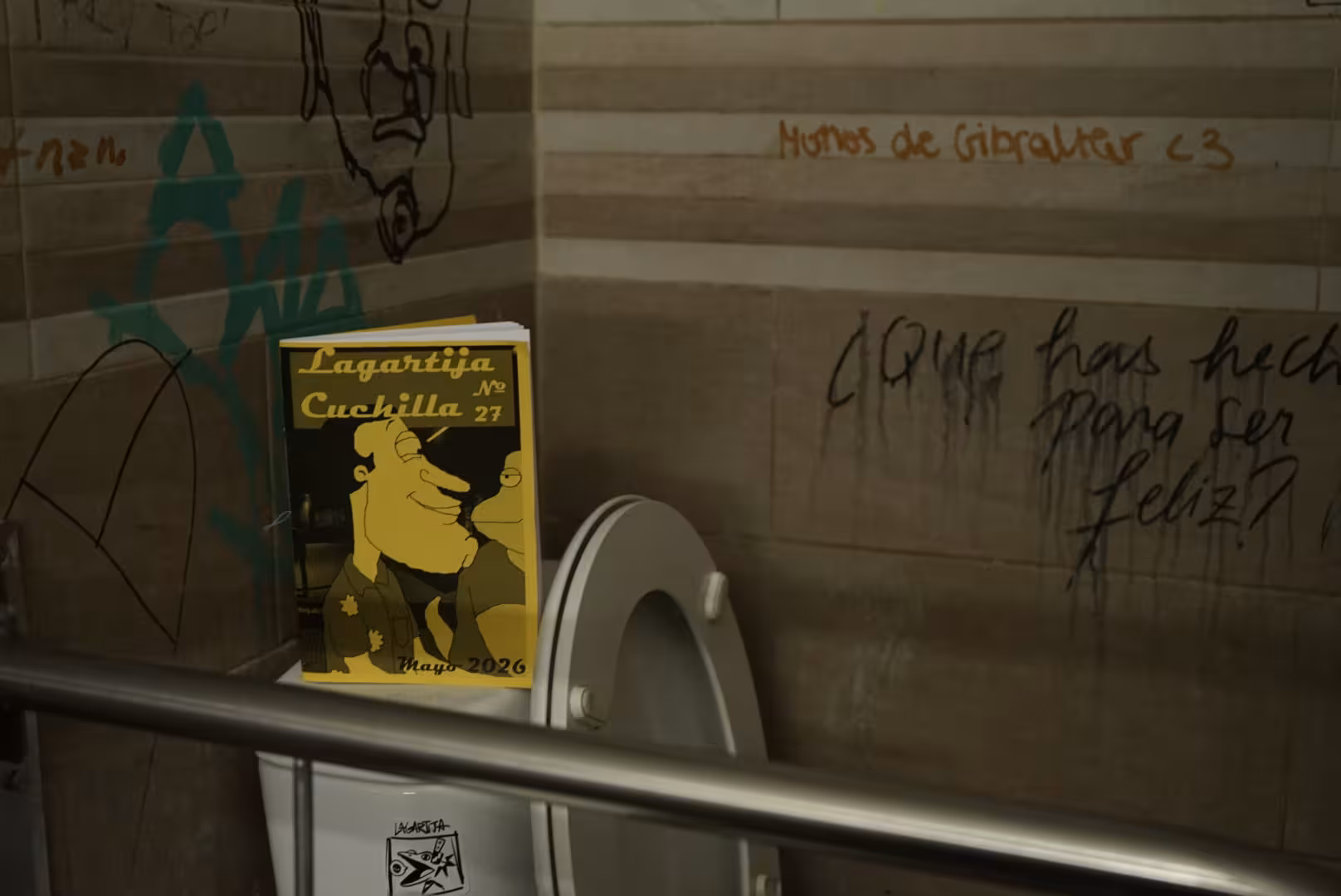 Foto del lanzamiento del número 26, a nuestra izquierda se puede ver la portada de este número, un poco más arriba una jaula vacía, al lado de la jaula un trapo verde, un dibujo de un personaje, y un dibujo de unas tazas y un vaso. A nuestra derecha se ve una silla azul vacía.